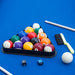 Full set of billiard accessories on blue felt, including racked multicolor balls, cue sticks, cue chalk, white cue ball, and table brush for maintenance.