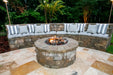 Luxury evening patio setup featuring the Travertine TessaRai 46" Round Gas Fire Pit with gas logs and a cushioned curved stone bench for outdoor entertaining.