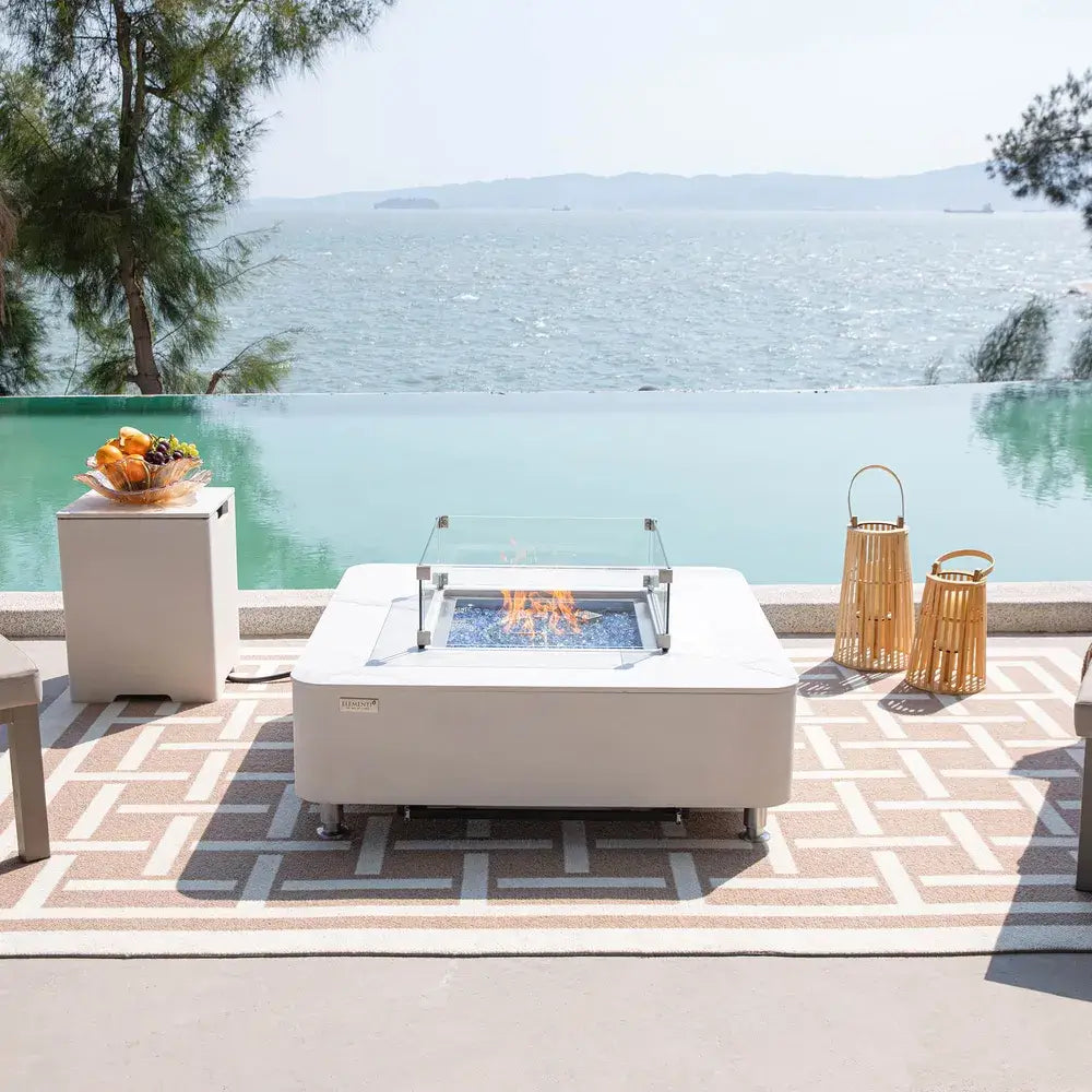 Luxury poolside patio featuring a burning Elementi Annecy fire table and matching white propane tank cover with a sea view.