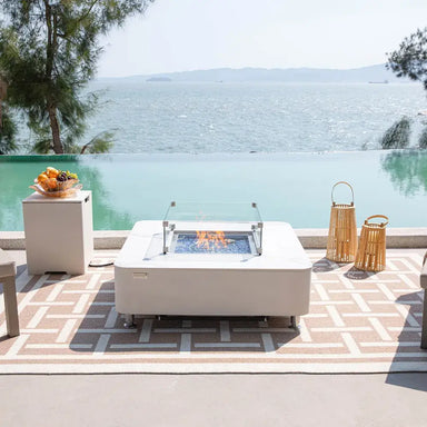 Luxury poolside patio featuring a burning Elementi Annecy fire table and matching white propane tank cover with a sea view.