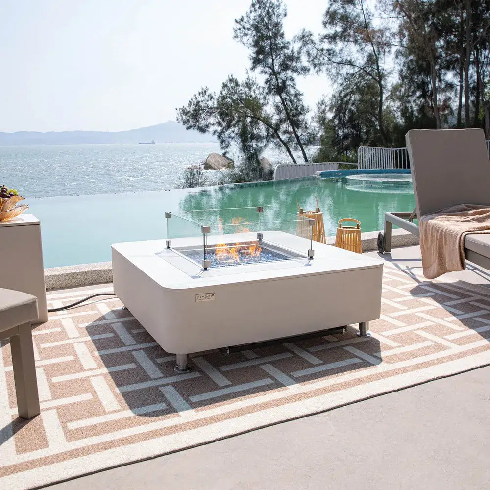 Modern outdoor terrace with a white porcelain Elementi fire pit, glass wind guard, and scenic waterfront backdrop.