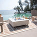Modern outdoor terrace with a white porcelain Elementi fire pit, glass wind guard, and scenic waterfront backdrop.