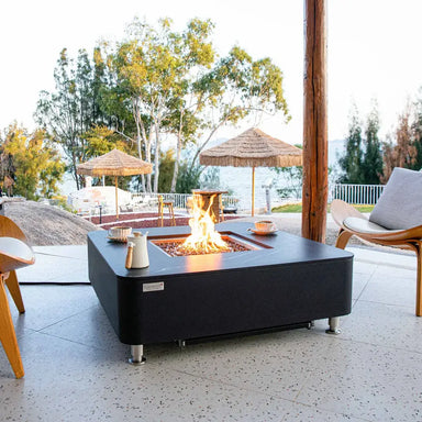 Modern outdoor patio setting with an Elementi OFP101BB fire table burning near a scenic waterfront view.