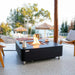 Modern outdoor patio setting with an Elementi OFP101BB fire table burning near a scenic waterfront view.