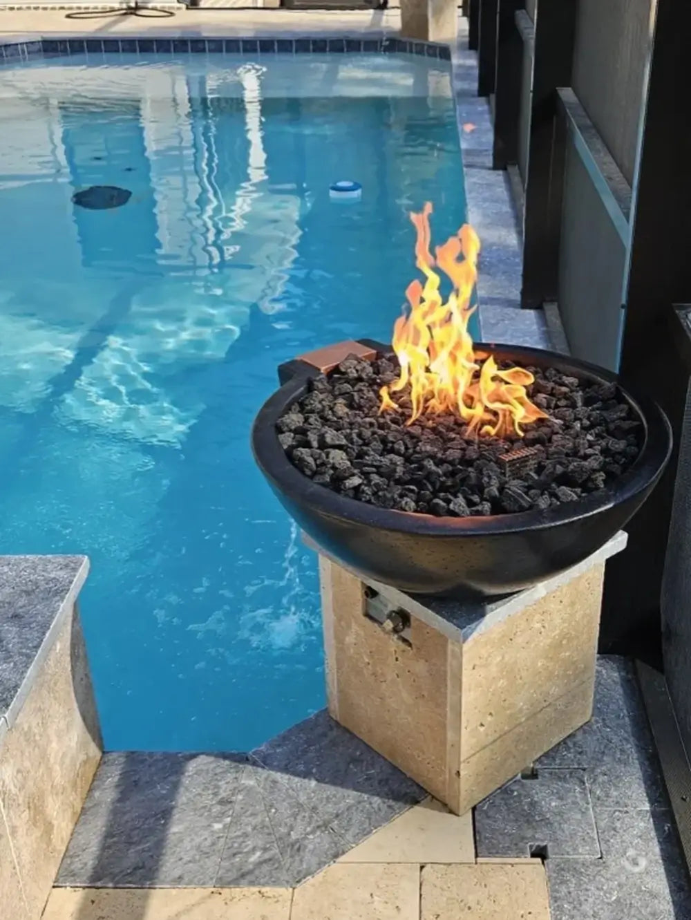 Black Travertine TessaRai fire and water bowl on pool pedestal with bright flames and modern backyard pool.