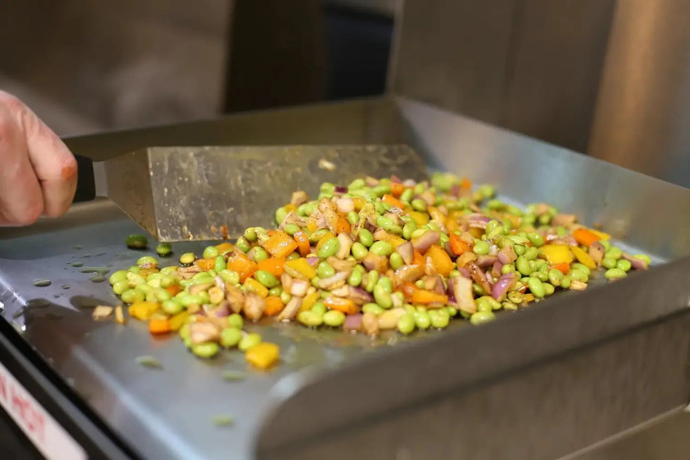 Colorful vegetables being stir-fried on Le Griddle flat-top cooktop surface.