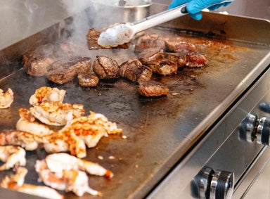 Chef grilling steak and chicken on stainless steel flat top griddle with even searing