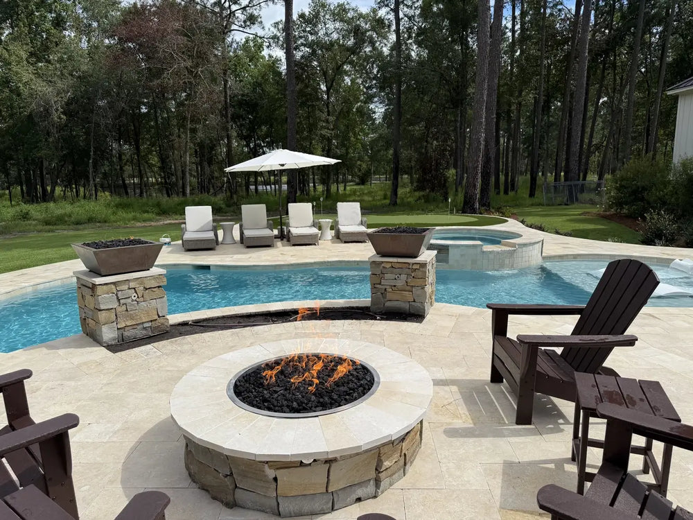 Poolside installation of the Travertine TessaRai 46" Round Gas Fire Pit surrounded by dark brown Adirondack chairs and matching stone fire pillars.
