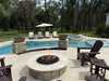 Poolside installation of the Travertine TessaRai 46" Round Gas Fire Pit surrounded by dark brown Adirondack chairs and matching stone fire pillars.