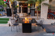 High-end outdoor dining area featuring the Brugge Bulgaria Black rectangular fire pit table, a designer woven chair, and a classic silver trailer in a backyard.