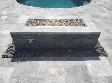 Outdoor stone fire pit featuring a Travertine T&M rectangle drop-in burner pan with gray fire glass, installed on a luxury silver travertine patio near a pool.