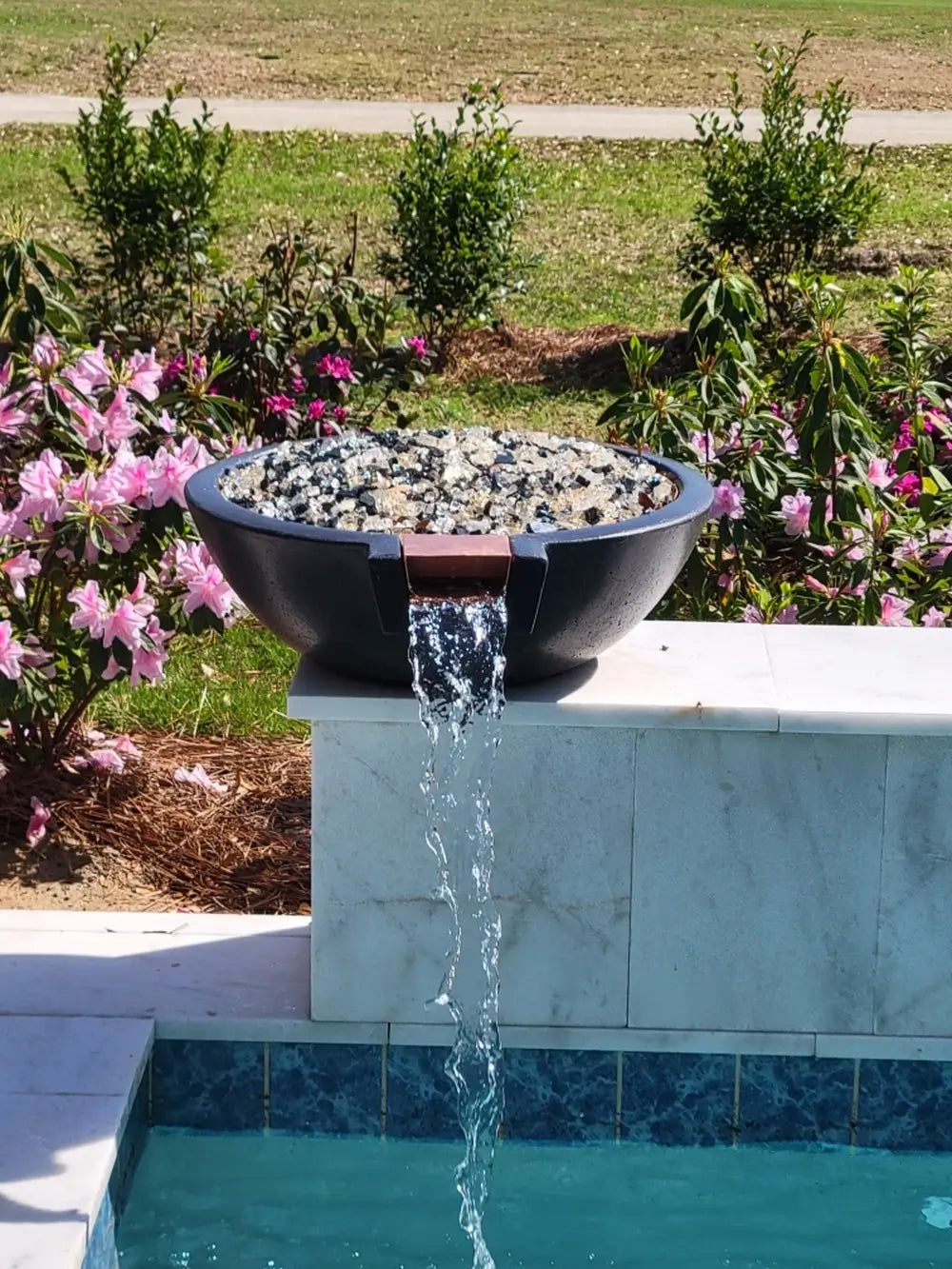 Travertine TessaRai fire and water bowl with clear fire glass and waterfall feature above resort-style pool.