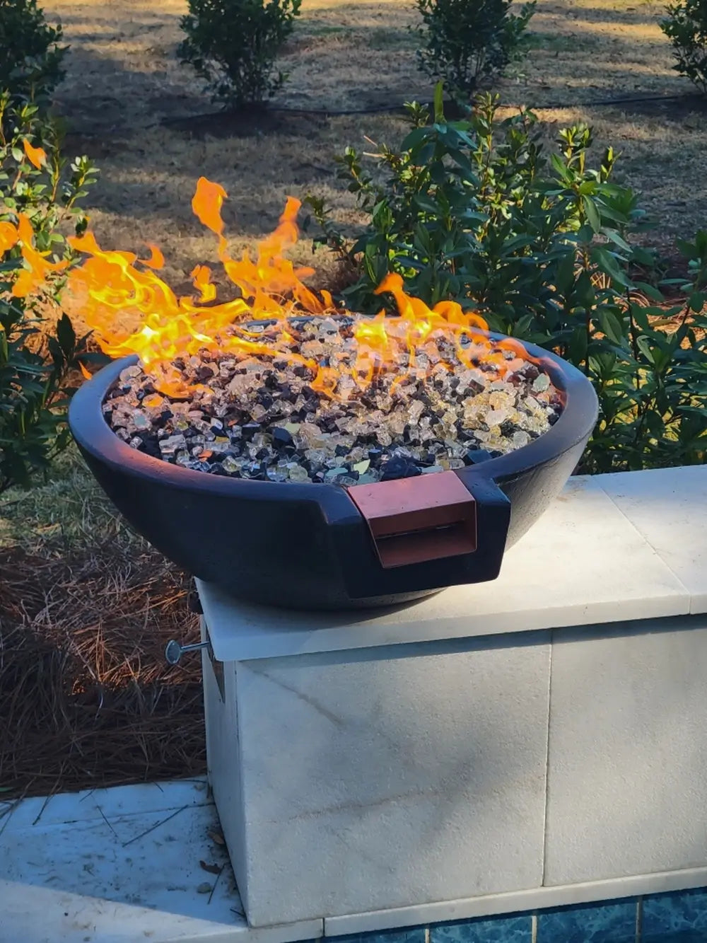 Black Travertine TessaRai fire and water bowl with fire glass on pool wall in landscaped backyard setting.