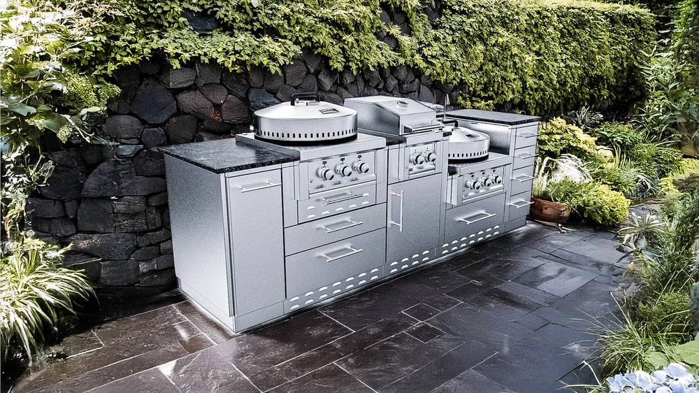 Close-up photograph of the Sunstone TexCajun outdoor kitchen island installed on a dark slate patio, showing its integrated burners and storage cabinets with a dark stone retaining wall and green vine background.