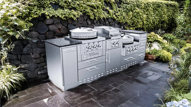 Close-up photograph of the Sunstone TexCajun outdoor kitchen island installed on a dark slate patio, showing its integrated burners and storage cabinets with a dark stone retaining wall and green vine background.