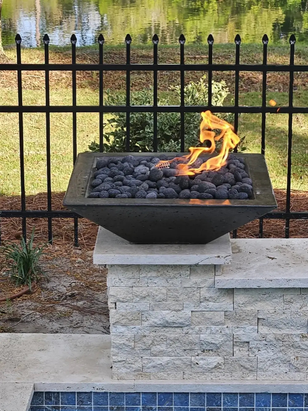 Black Travertine TessaRai 36 inch Square Fire Bowl Kit with lava rock and flame beside fence and waterfront yard