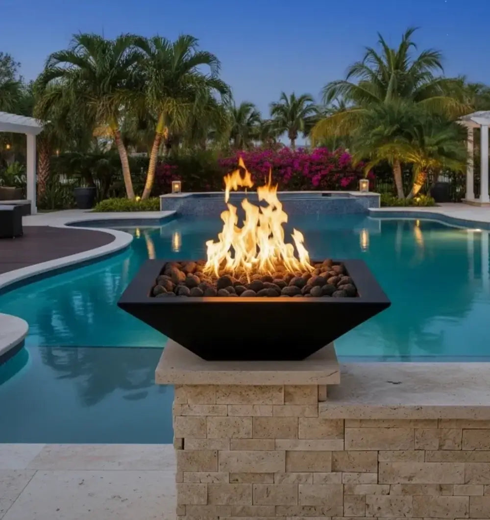 Travertine TessaRai 36 inch Square Fire Bowl Kit glowing at dusk beside luxury swimming pool and tropical landscaping