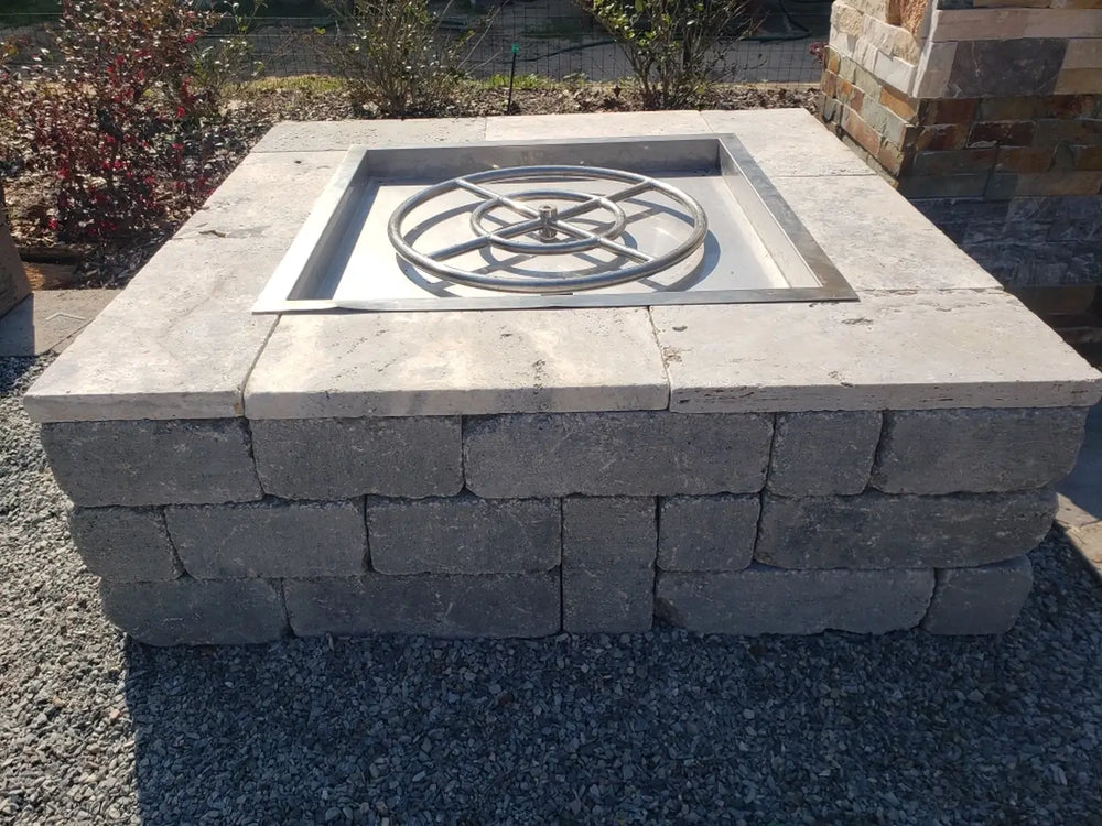 TessaRai square gas fire pit kit under construction featuring a grey stone block base and light travertine top caps with a stainless steel burner insert.