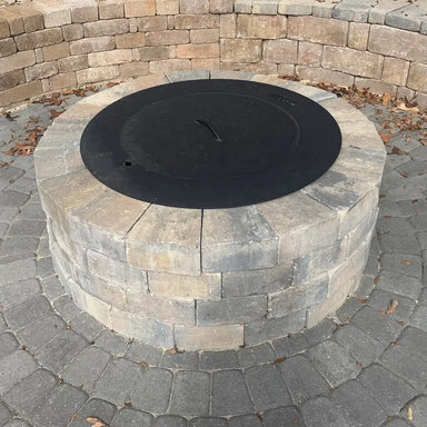 Round stone fire pit with a black protective lid cover installed on a circular paver patio.