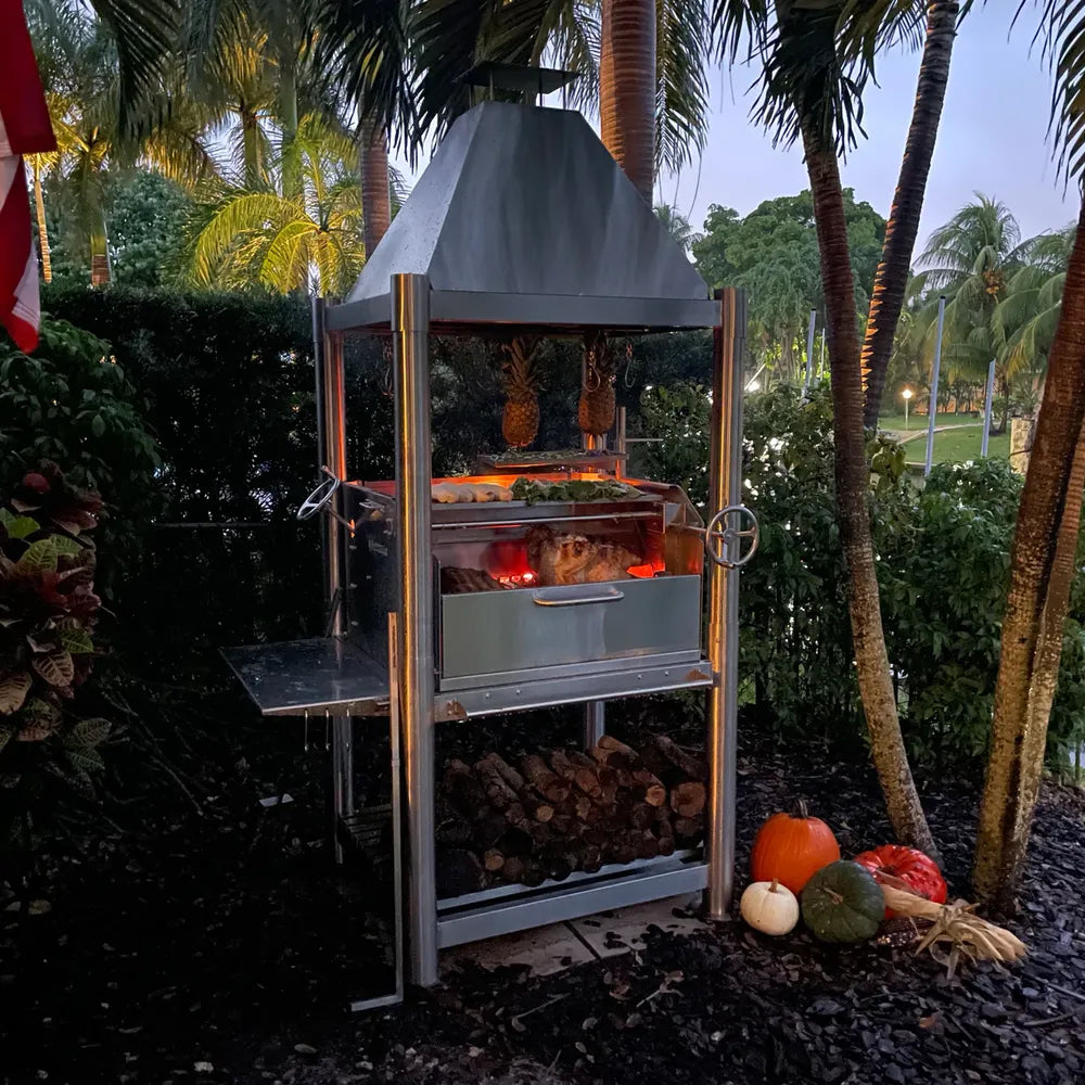 Tagwood BBQ BBQ02SS SS304 grill at dusk with wood fire and pumpkins by palm trees