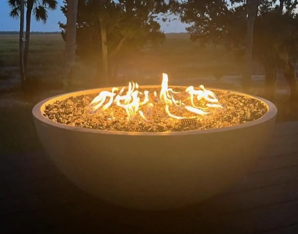 Travertine TessaRai 40 inch Patio Fire Pit Kit glowing at dusk with reflective fire glass on a scenic backyard deck