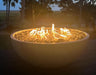 Travertine TessaRai 40 inch Patio Fire Pit Kit glowing at dusk with reflective fire glass on a scenic backyard deck