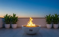 Travertine TessaRai 40 inch Patio Fire Pit Kit with tall flames on a modern rooftop terrace framed by potted tropical plants at dusk