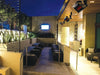 Multiple Bromic Platinum series heaters mounted on a high ceiling above a contemporary, sunken outdoor patio area with a television and modern seating.