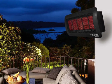 The Bromic Tungsten 300 Gas Radiant Heater mounted on a wall above a patio seating area, overlooking a beautiful night-time view of a bay with boats.