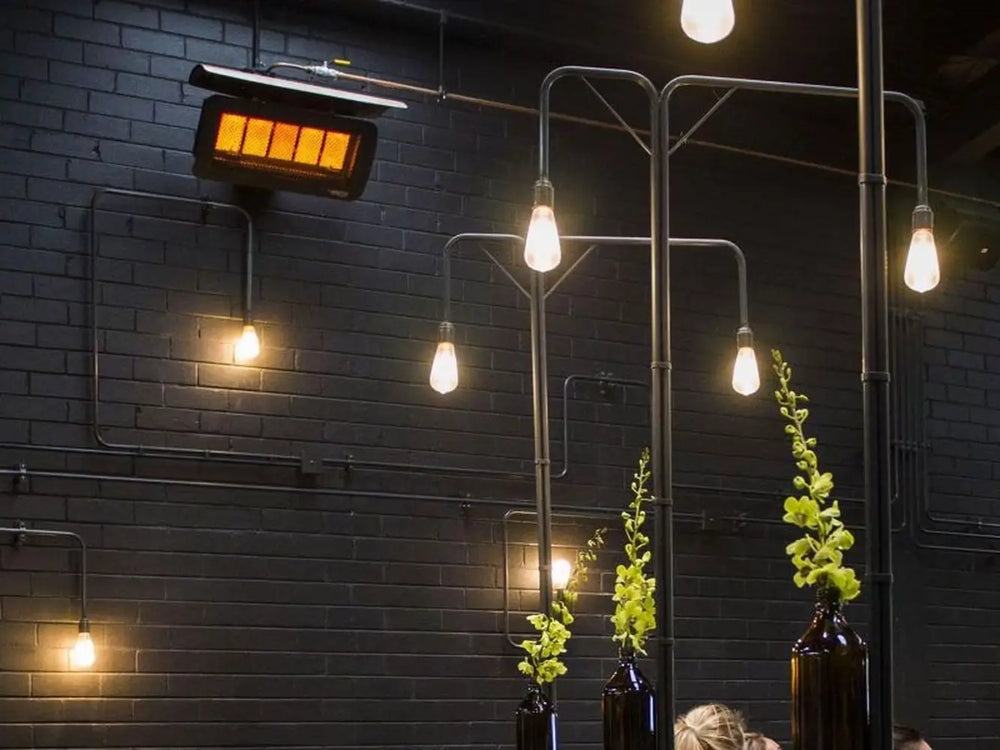 The Bromic Tungsten 300 Gas Radiant Heater mounted on an exposed pipe against a black brick wall, blending into a contemporary restaurant or bar environment.