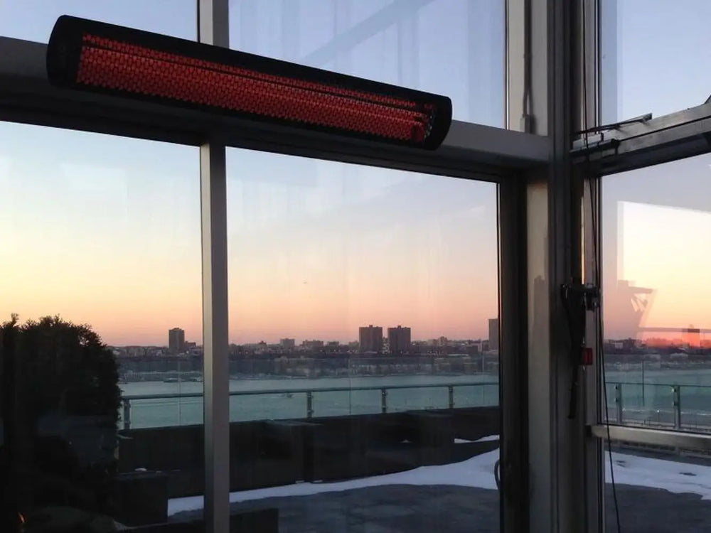 Bromic Smart-Heat black electric heater wall-mounted by large windows overlooking a city skyline at sunset.