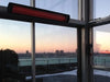 Bromic Smart-Heat black electric heater wall-mounted by large windows overlooking a city skyline at sunset.