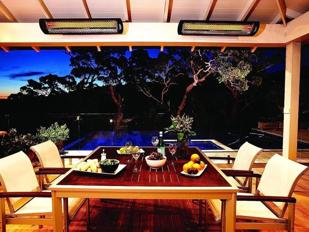 Two Bromic Tungsten 6000W Electric Heaters in black installed above a wooden patio dining area, illuminating a cozy evening meal setup near a pool.