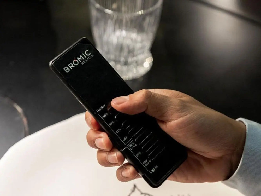 Hand holding the black Bromic Eclipse Dimmer Controller remote, showing 3-stage heat and light adjustment buttons.