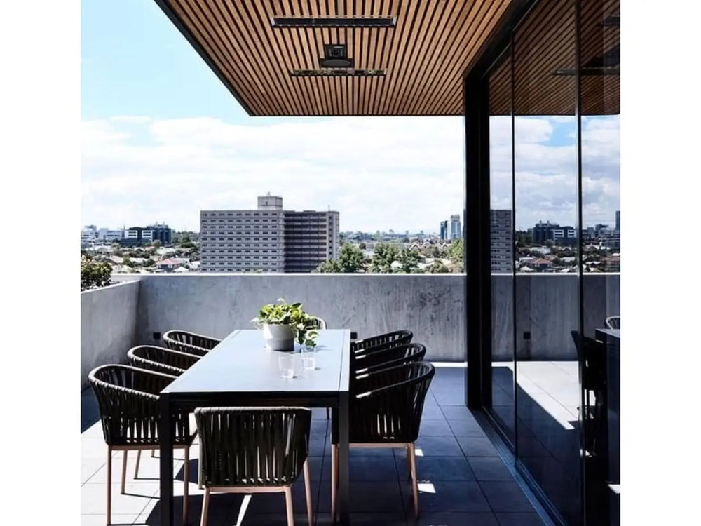 The Bromic Platinum 4500W Electric Heater mounted on a wooden-slat ceiling, providing heat to a chic outdoor patio with a dining table and a cityscape view.