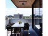 The Bromic Platinum 4500W Electric Heater mounted on a wooden-slat ceiling, providing heat to a chic outdoor patio with a dining table and a cityscape view.