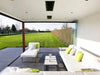 The Bromic Platinum 4500W Electric Heater recessed into a ceiling, blending seamlessly into a luxurious, all-white outdoor lounge area with a large lawn and hedge in the background.