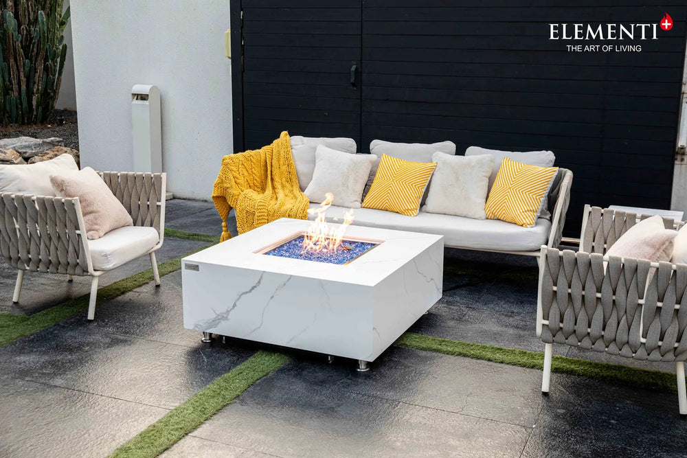 Elementi Plus OFP103BW Bianco 60,000 BTU fire table featuring a white porcelain marble-effect top, styled on a modern patio with black outdoor sofas and green artificial turf.