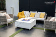 Elementi Plus OFP103BW Bianco 60,000 BTU fire table featuring a white porcelain marble-effect top, styled on a modern patio with black outdoor sofas and green artificial turf.