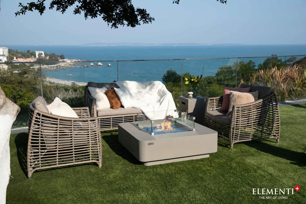 Elementi Plus Lucerne fire table with glass wind guard on terrace with ocean view and wicker seating.