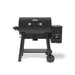 Front view of the Broil King Crown Pellet 500 WiFi pellet smoker in matte black, featuring a barrel-style cooking chamber, chrome handle, side pellet hopper with digital controller, small front shelf, lower support shelf, and two transport wheels on a light gray background.