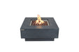 Square Elementi Manhattan fire pit table in dark grey concrete with a center flame and red lava rocks on a white background.