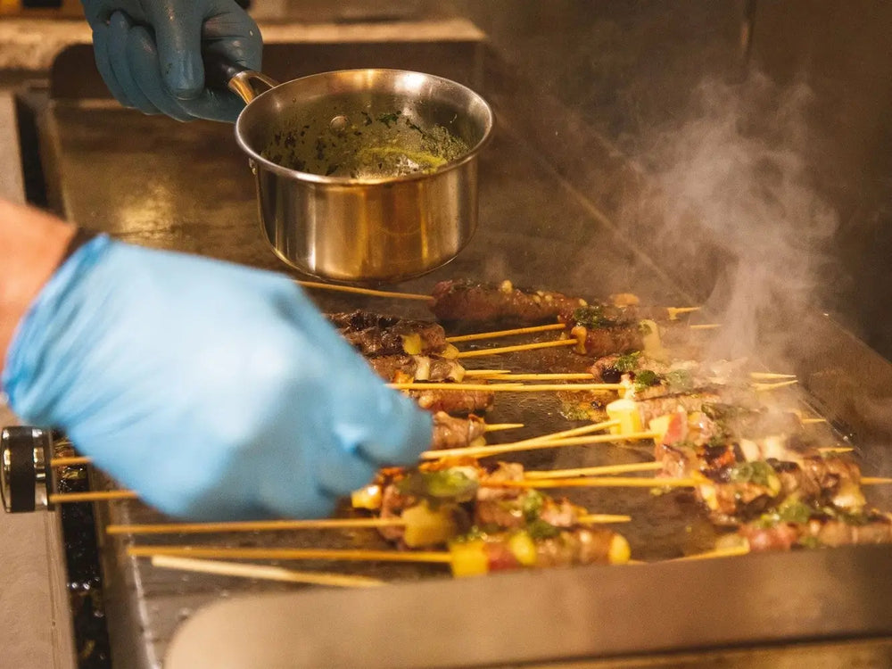 Close-up of skewers cooking on a Le Griddle, with sauce being poured from a small pot.