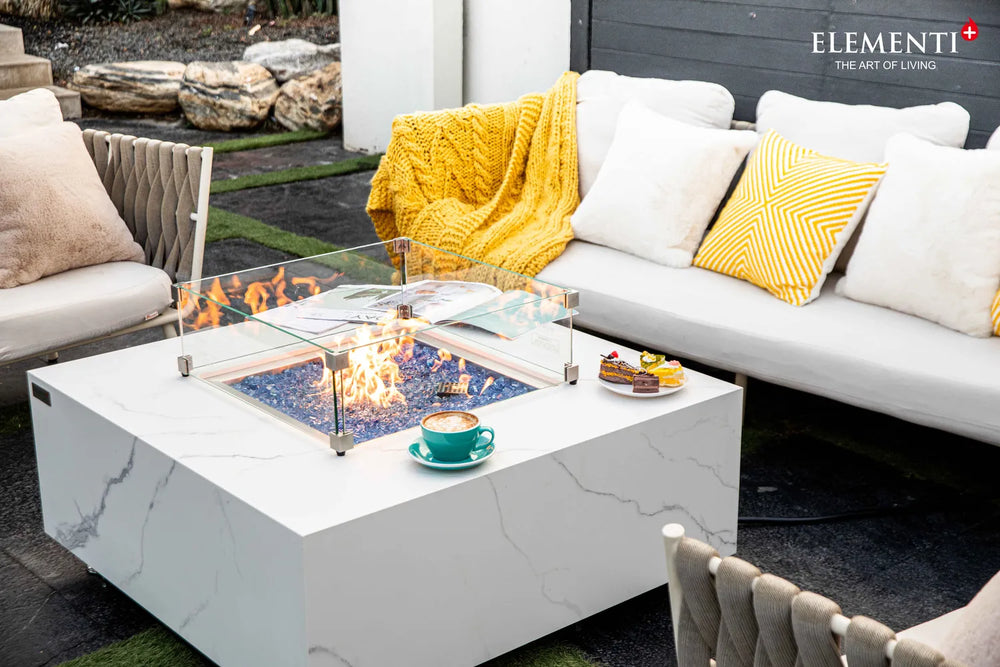 Modern backyard patio scene showcasing the Elementi Plus Bianco rectangular gas fire pit table as a centerpiece for a luxury outdoor lounge area.
