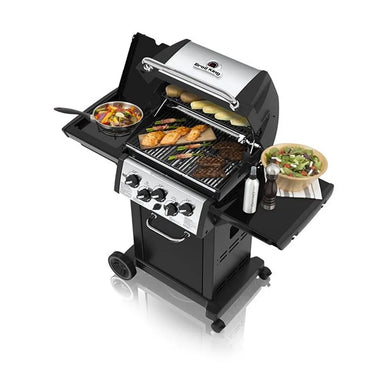 Broil King Monarch 390 gas grill with lid open and food cooking on the grates, featuring an integrated side burner with skillet, warming rack, black shelves, and portable cart design