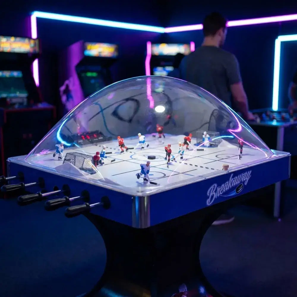 Hathaway Breakaway Dome Hockey table shown in an arcade setting with neon lights, focusing on the clear dome and blue table sides.