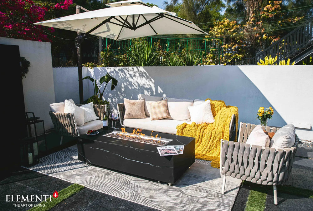 Modern outdoor living area featuring the Varna Bulgaria Black square gas fire pit table as a centerpiece, paired with a designer woven chair and a vintage silver trailer.