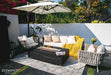 Modern outdoor living area featuring the Varna Bulgaria Black square gas fire pit table as a centerpiece, paired with a designer woven chair and a vintage silver trailer.